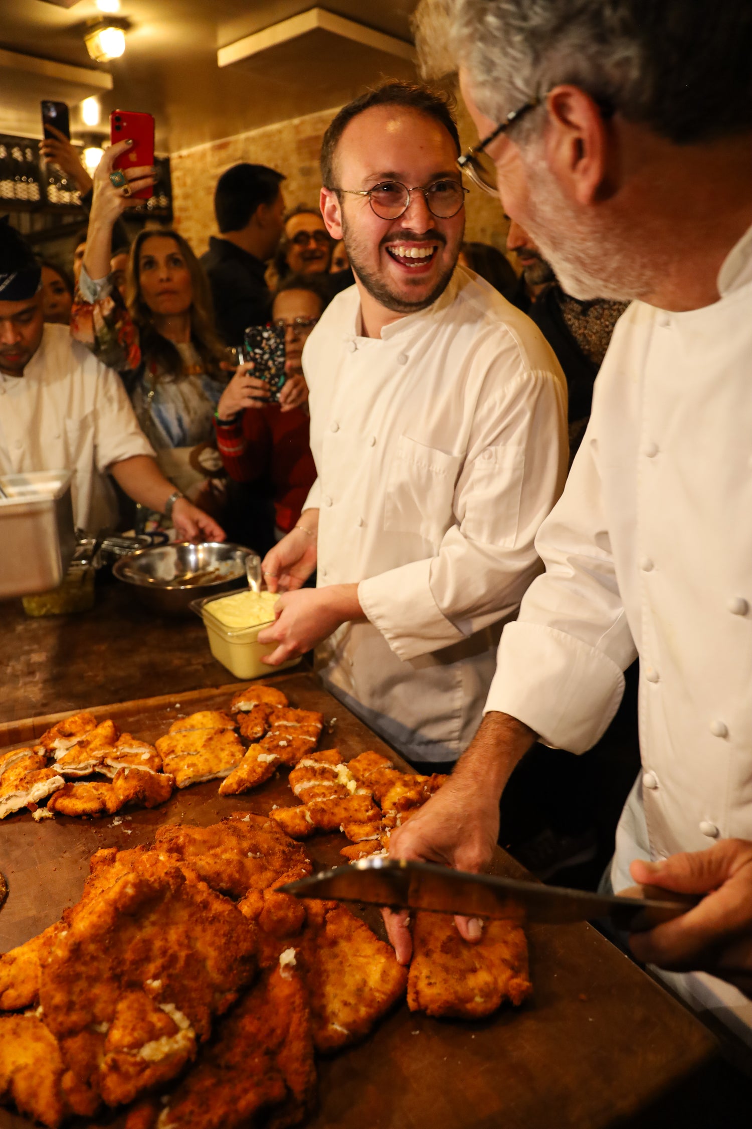 A warm, bustling restaurant scene with two chefs in white uniforms serving golden-brown fried chicken from a large wooden platter. One chef is smiling broadly while the other slices the food, with other diners and staff visible in the background.