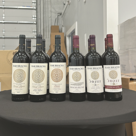 A selection of Har Bracha wines displayed on a table, featuring bottles of Highlander Special Reserve Merlot and Jozef Syrah Merlot, with a blurred warehouse background.