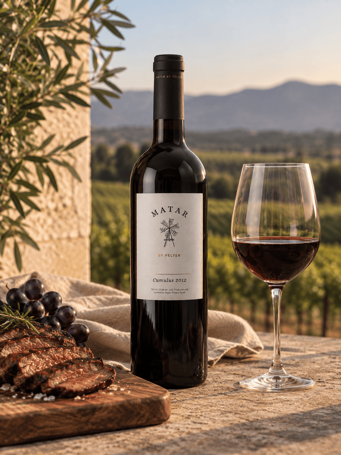 A bottle of Matar Cumulus 2023 red wine stands on a stone table beside a filled wine glass, with sliced steak, grapes, and a vineyard landscape in the background.