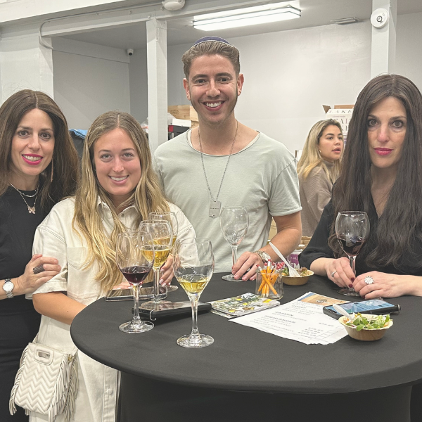 Three guests are smiling at a wine tasting event, each holding a glass of wine, with appetizers on the table in front of them and event attendees in the background.