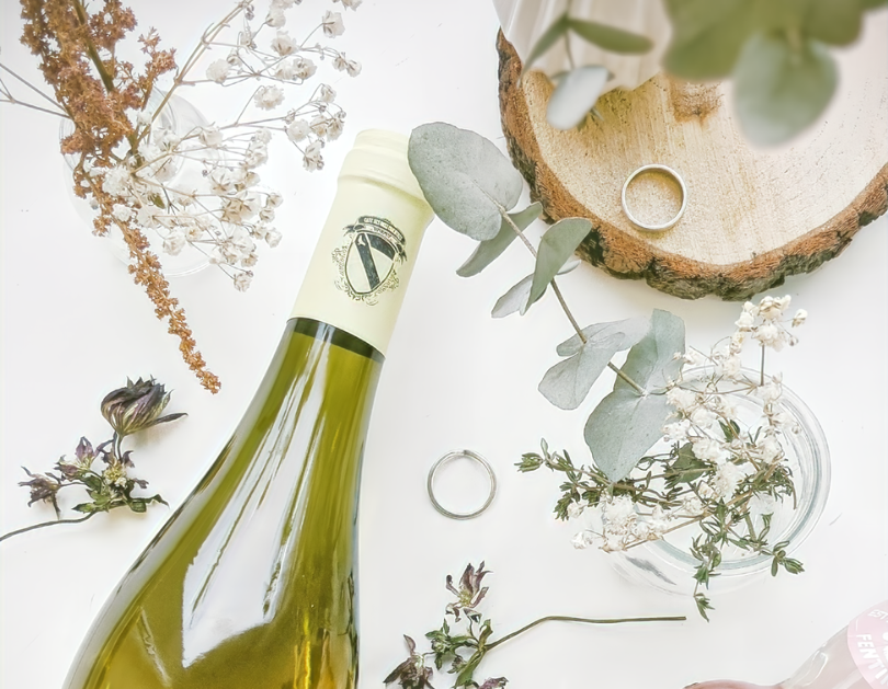 Wine bottle surrounded by flowers, leaves, and wedding rings.