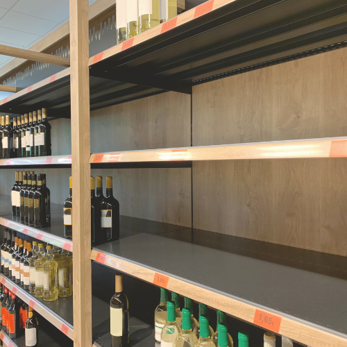 A wine store shelf partially stocked with bottles, with noticeable gaps indicating a shortage, and price tags visible on the shelf edges.