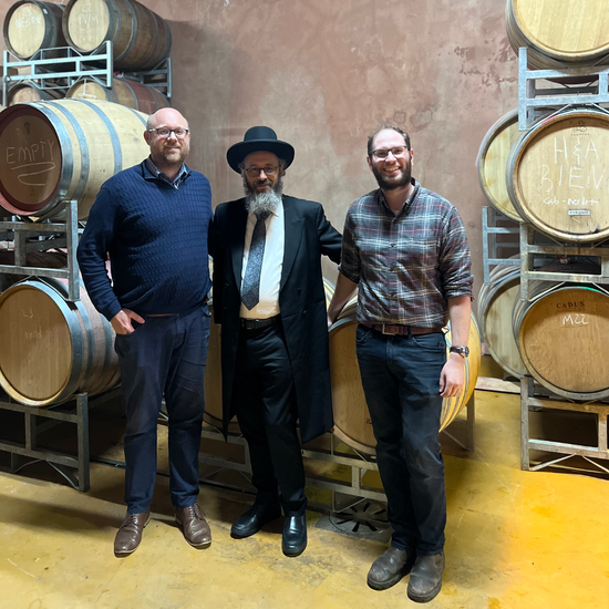 Three men standing in a winery or distillery, surrounded by wooden barrels stacked on metal racks