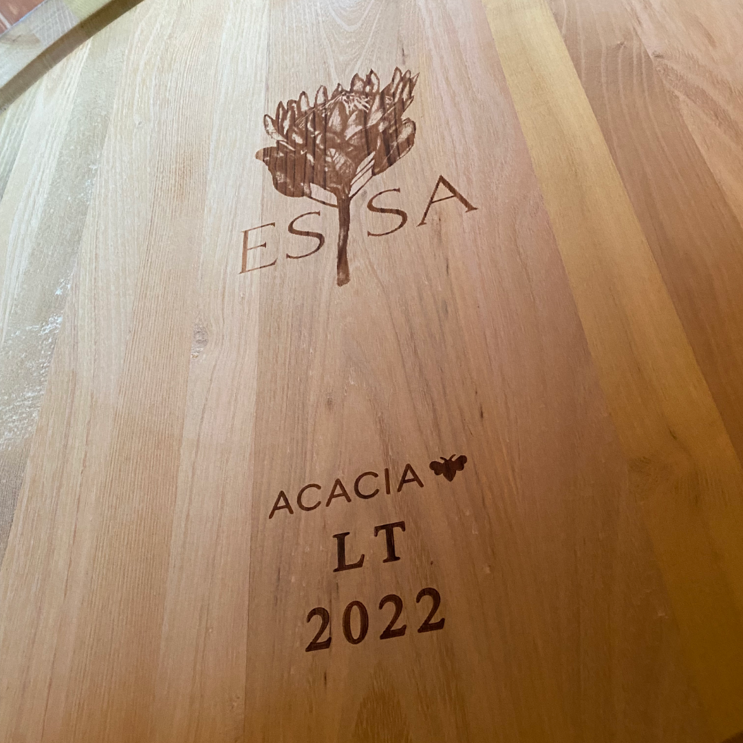 A close-up of a wooden surface with engravings. The engravings include a detailed illustration of a flower or plant above the word "ESSA."