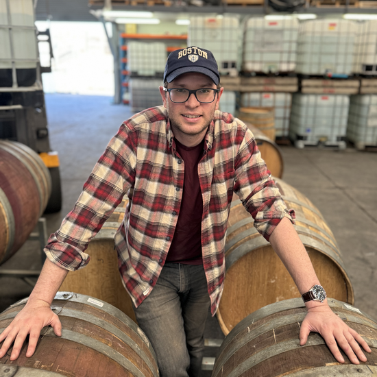 A man wearing a plaid shirt, a Boston cap, and glasses, leaning on wooden barrels inside a winery or distillery