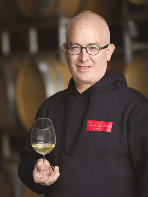 Amichai Lourie, wearing glasses and a black hoodie with a red "Shiloh Winery" label, holds a glass of white wine, standing in front of wine barrels.