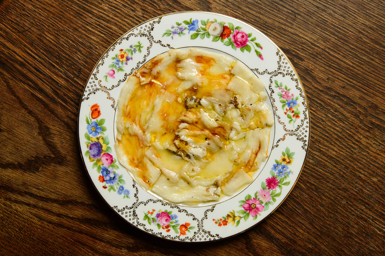 A top-down view of eggplant carpaccio served on a white ceramic plate with a colorful floral border, set on a dark wooden table.
