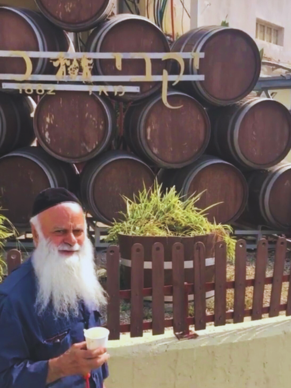 A man with a white beard, wearing a black cap and holding a cup, standing in front of a stack of large wooden barrels at Carmel Winery.