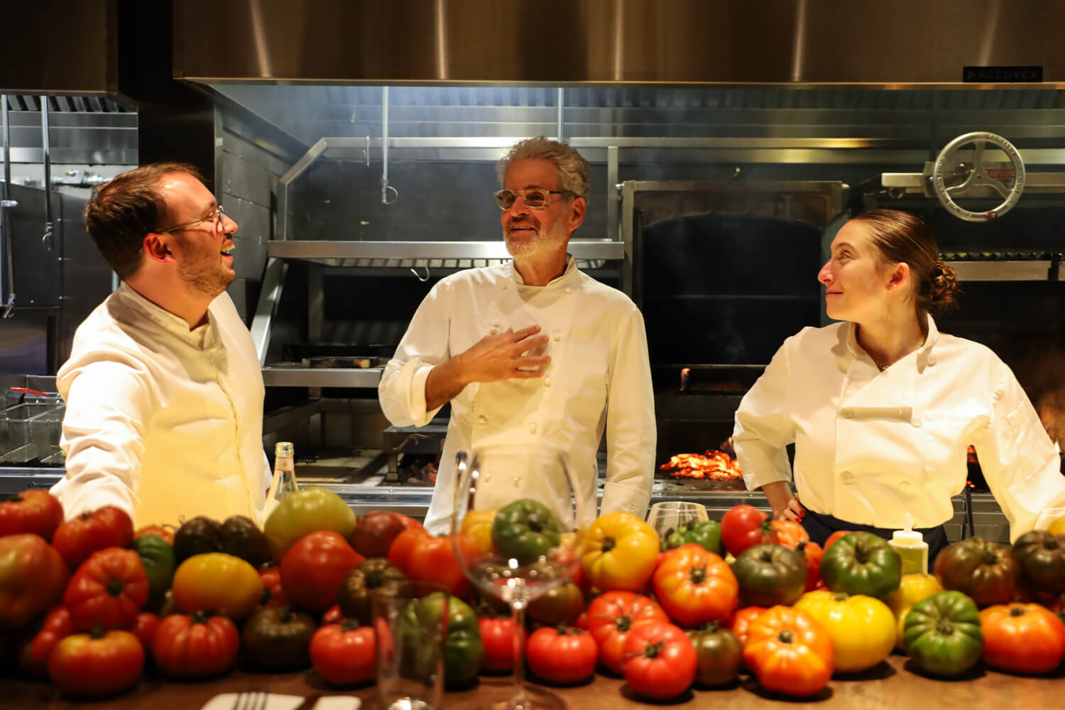Three chefs in white uniforms standing in a professional kitchen, with a large assortment of colorful heirloom tomatoes arranged on a counter in the foreground.