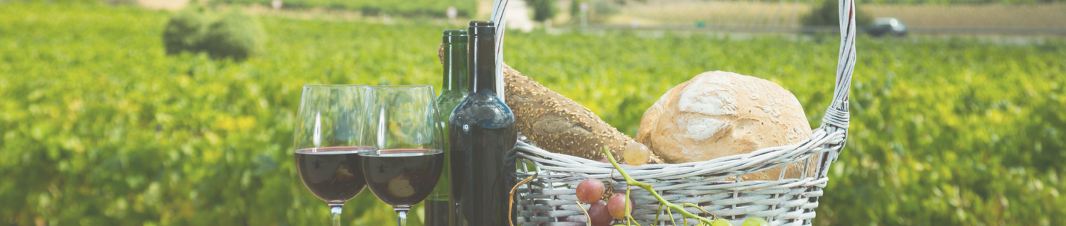 A vineyard background with a picnic basket containing wine bottles, glasses of red wine, grapes, and bread, representing a wine and food celebration.