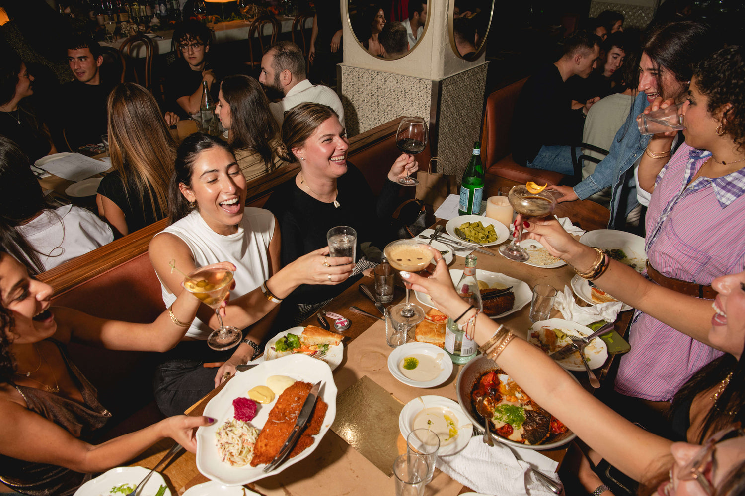 A group of friends laughing and toasting around a dining table filled with food and drinks in a restaurant setting.