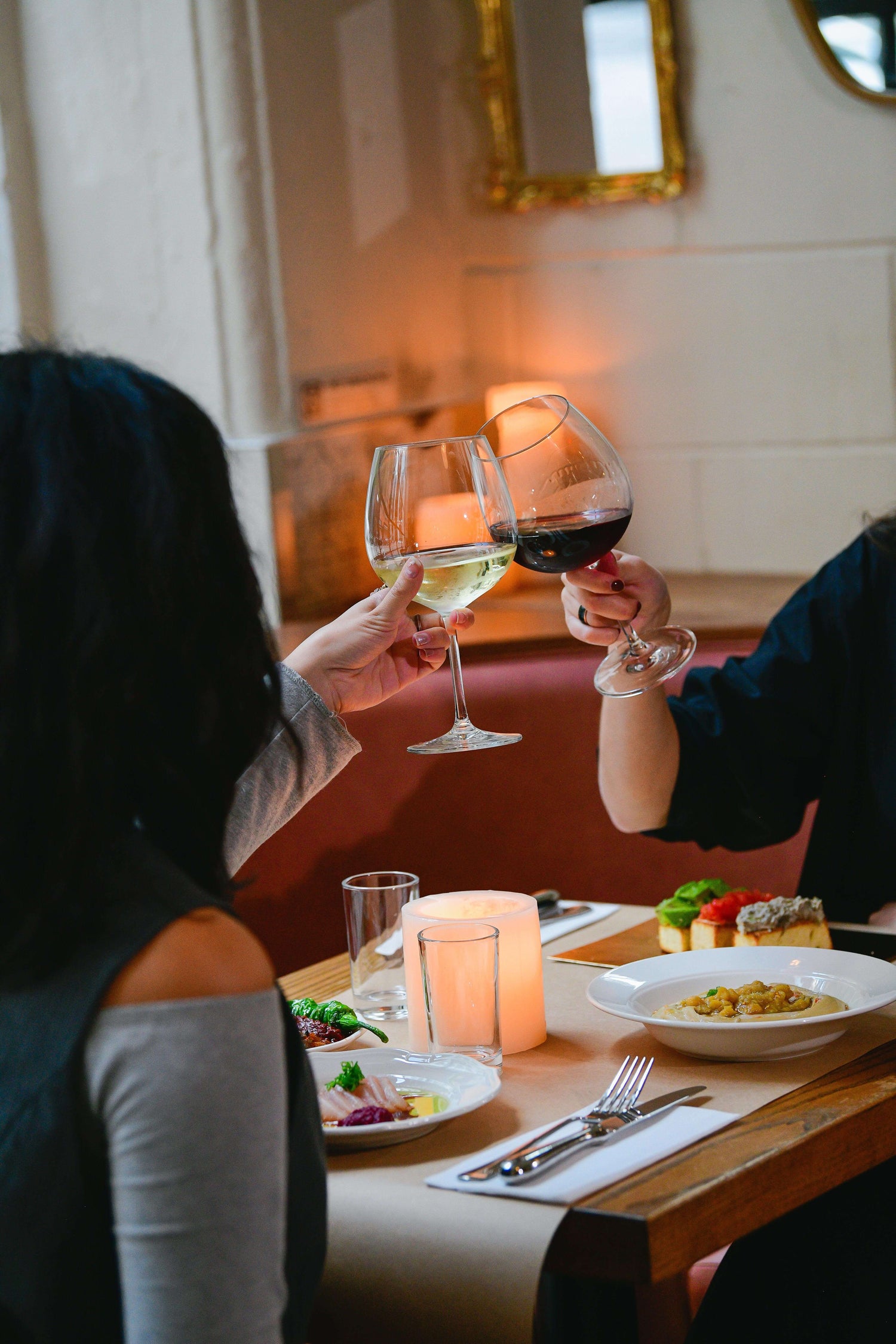 A cozy restaurant dinner scene with two people raising wine glasses in a toast, featuring red and white wine, candlelight, plated food, and a warm, intimate dining atmosphere.
