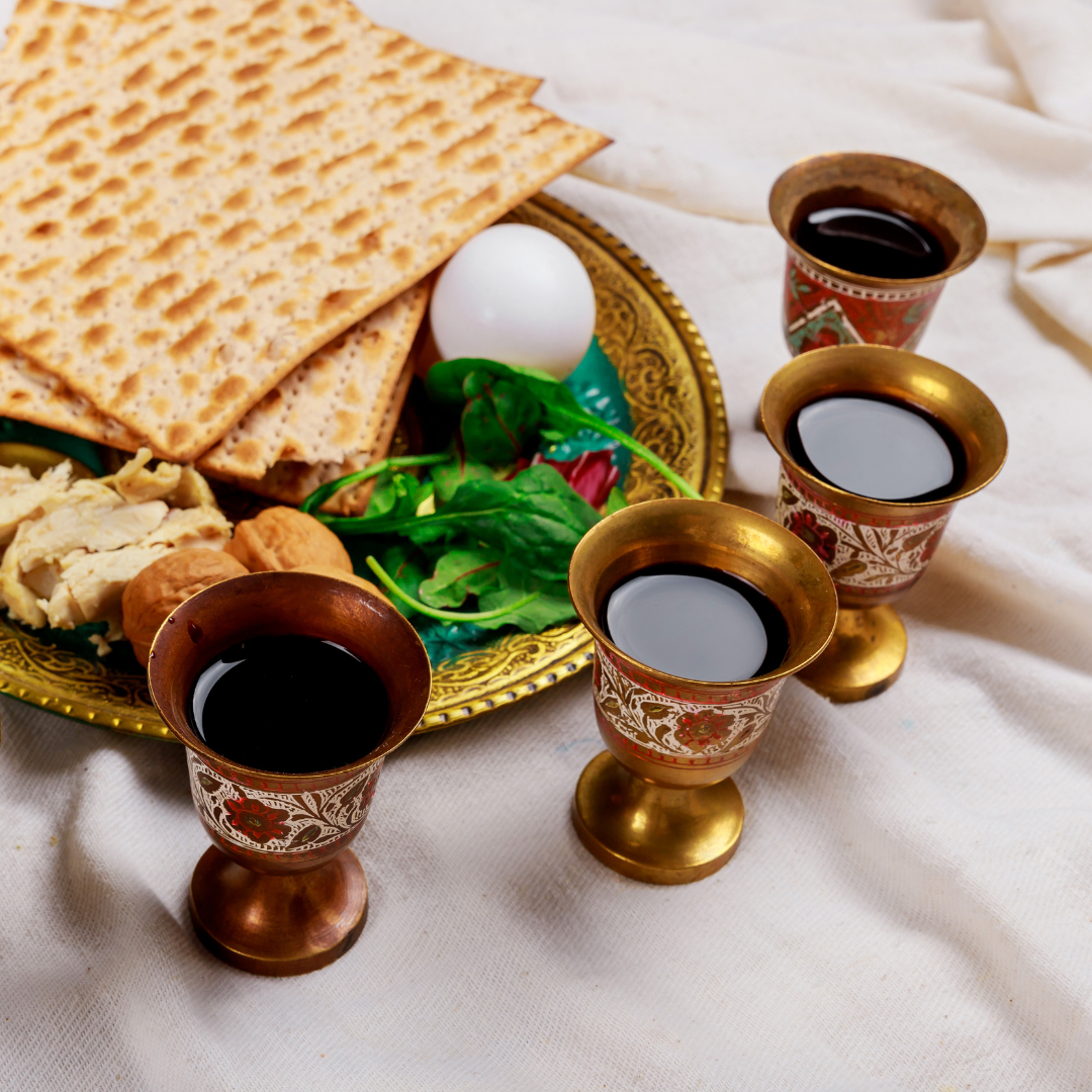 A traditional Seder plate with matzo, an egg, and bitter herbs accompanied by three ornate wine goblets filled with red wine.