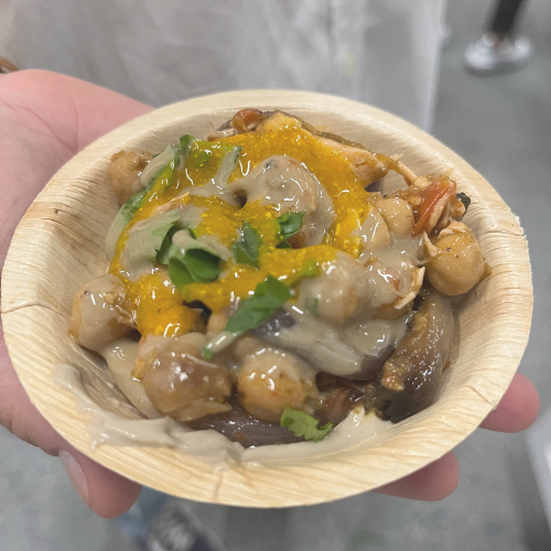 A hand holds a bowl with a savory dish featuring chickpeas, eggplant, and a drizzle of yellow sauce.