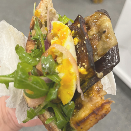 A close-up view of a hand holding a sandwich with grilled eggplant, arugula, and a hard-boiled egg.