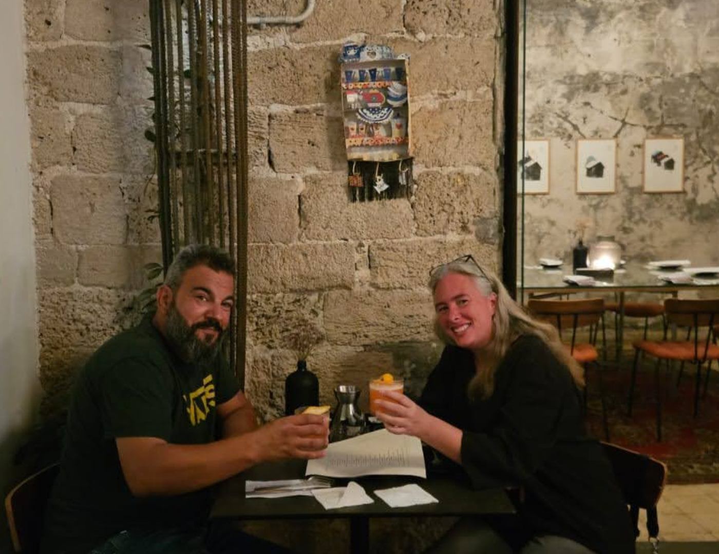 Two people sitting at a table in a rustic setting with stone walls and a small display of bottles.
