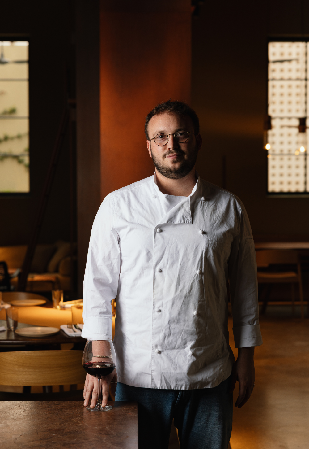 Chef Loel Miller wearing a white double-breasted chef coat and glasses stands in a dimly lit restaurant or kitchen environment, holding a glass of red wine.