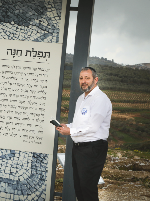 A man in a white shirt and black pants stands next to a large prayer plaque. He holds a prayer book, with vineyards visible in the background under a cloudy sky.