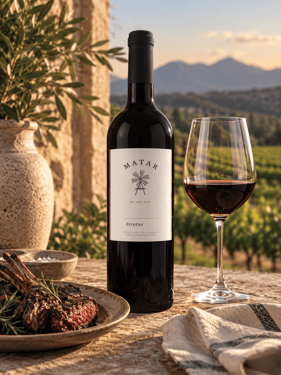 A bottle of Matar Stratus red wine stands on a stone table beside a filled wine glass, with a plate of lamb and a vase in the foreground against a vineyard and mountain landscape at sunset.