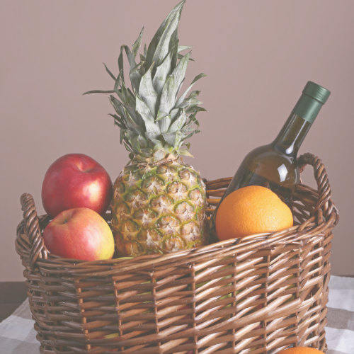 A wicker basket containing a pineapple, apples, an orange, and a bottle of white wine, suggesting a celebratory or gift setting.