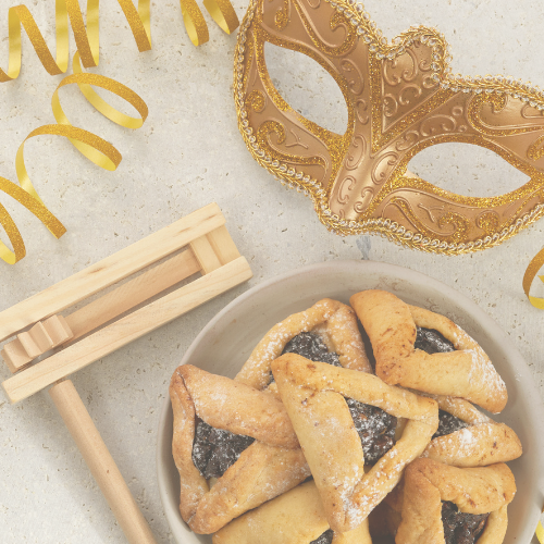 A festive Purim setup featuring a golden mask, hamantaschen cookies in a bowl, and a wooden gragger, with golden streamers on a textured surface.