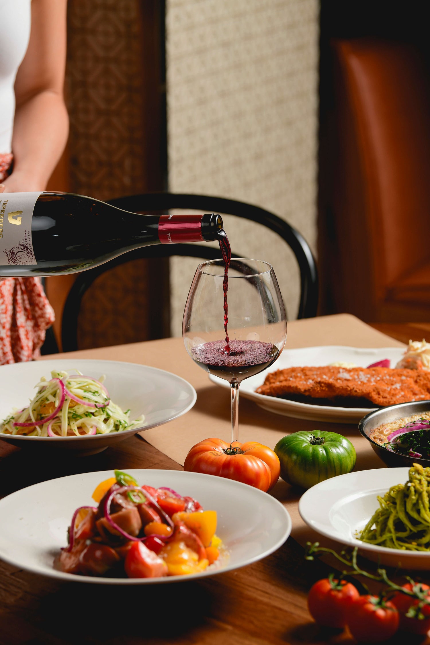 A warm restaurant dining scene featuring red wine being poured into a wine glass, surrounded by colorful gourmet dishes, fresh vegetables, and tomatoes on a wooden table, with cozy ambient lighting and an intimate atmosphere.
