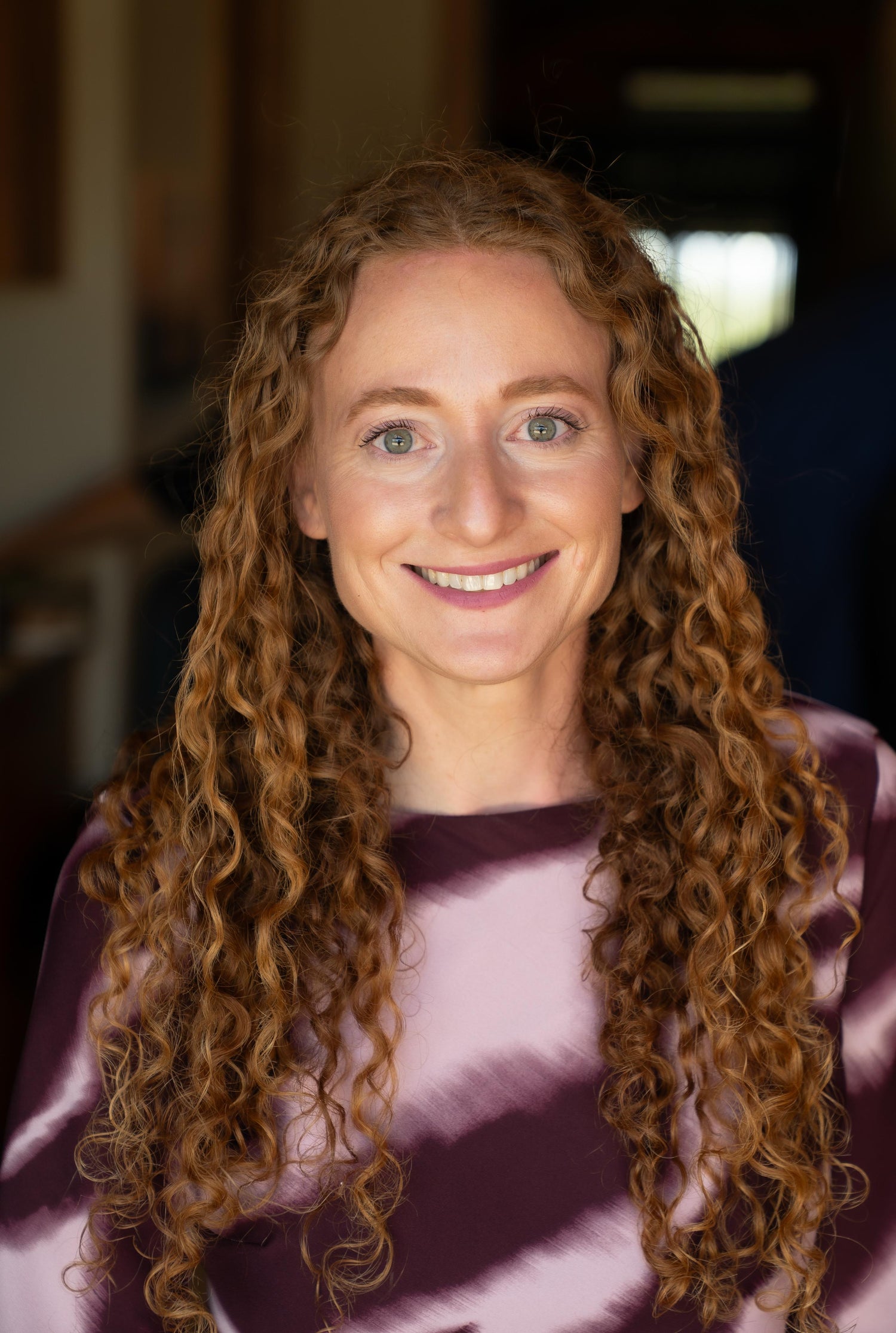 Rochel Raitman  with long, curly light brown hair wearing a purple and pink tie-dye top, photographed in a softly blurred indoor setting.