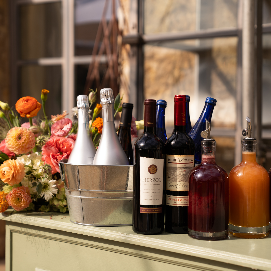 Display of kosher wine and beverages featuring sparkling wine, red wine bottles, and colorful drink dispensers beside a vibrant floral arrangement on a rustic table at an elegant event venue.