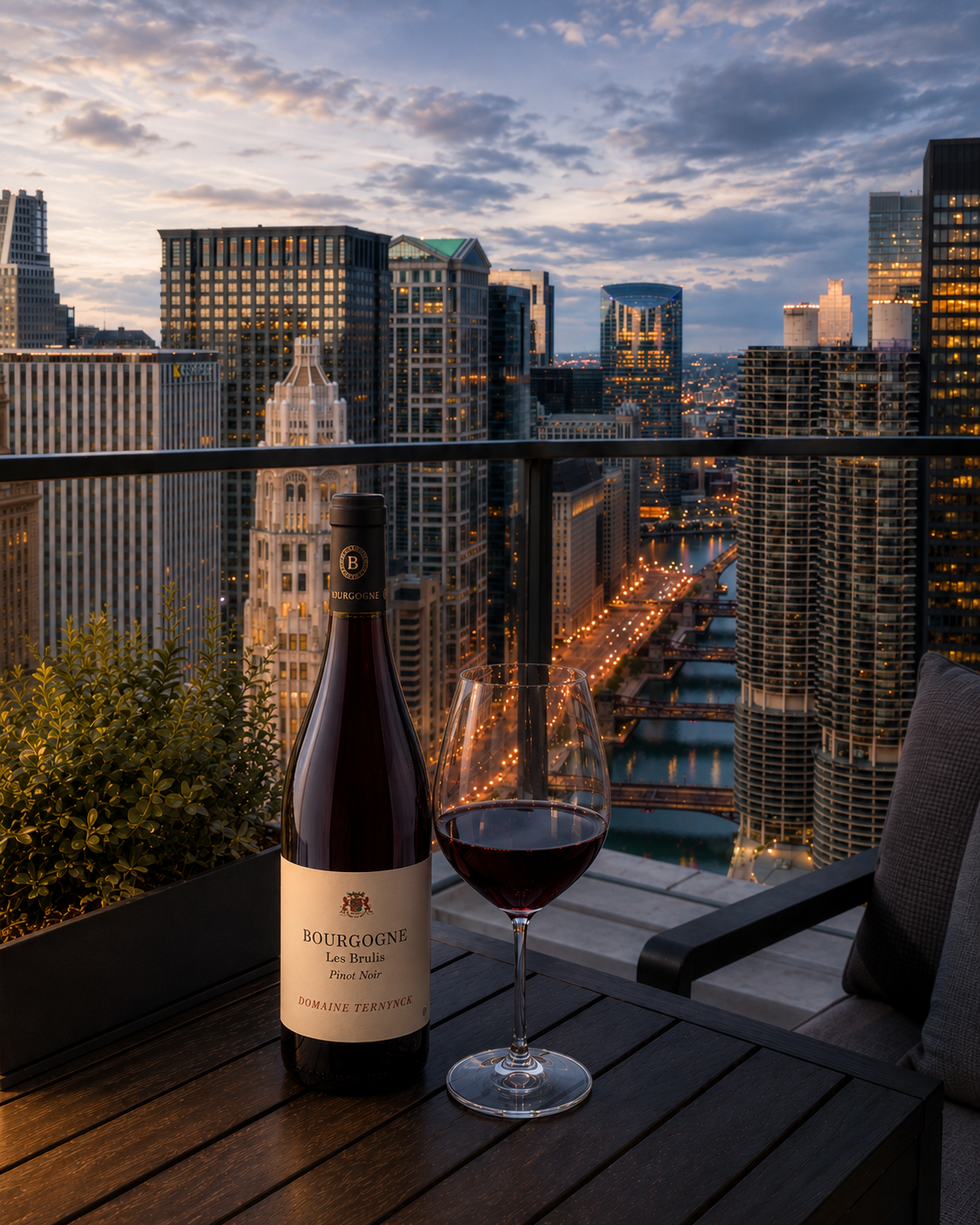 Elegant Chicago rooftop terrace at dusk featuring a bottle of Bourgogne Pinot Noir and a glass of red wine on a dark wood table, with the Chicago skyline and river visible in the background.