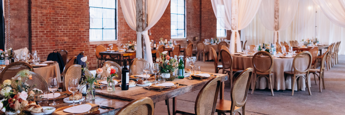 A large banquet hall or event space with brick walls, tall windows, draped white fabric, and multiple round tables set with linens, glassware, and floral centerpieces.