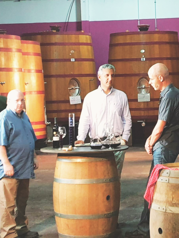 Three men standing around a wine barrel table with wine glasses and bottles, inside a winery with large wooden vats in the background.