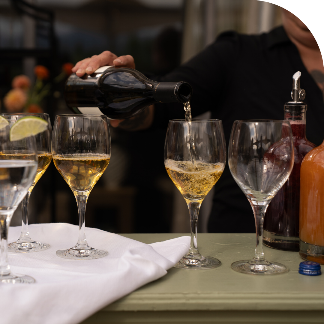 A server pours white wine into a glass at an event bar, with several wine glasses, fresh lime garnishes, and bottles of kosher wine and handcrafted beverages arranged on the counter.